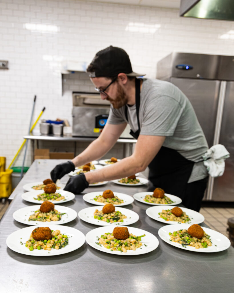 A cook in a black cap and gloves plating a row of dishes in the Cinderlands Warehouse kitchen