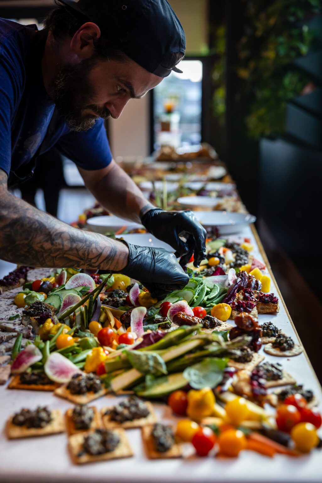 A Cinderlands chef in a cap and gloves arranging vegetables and crackers on a long grazing board