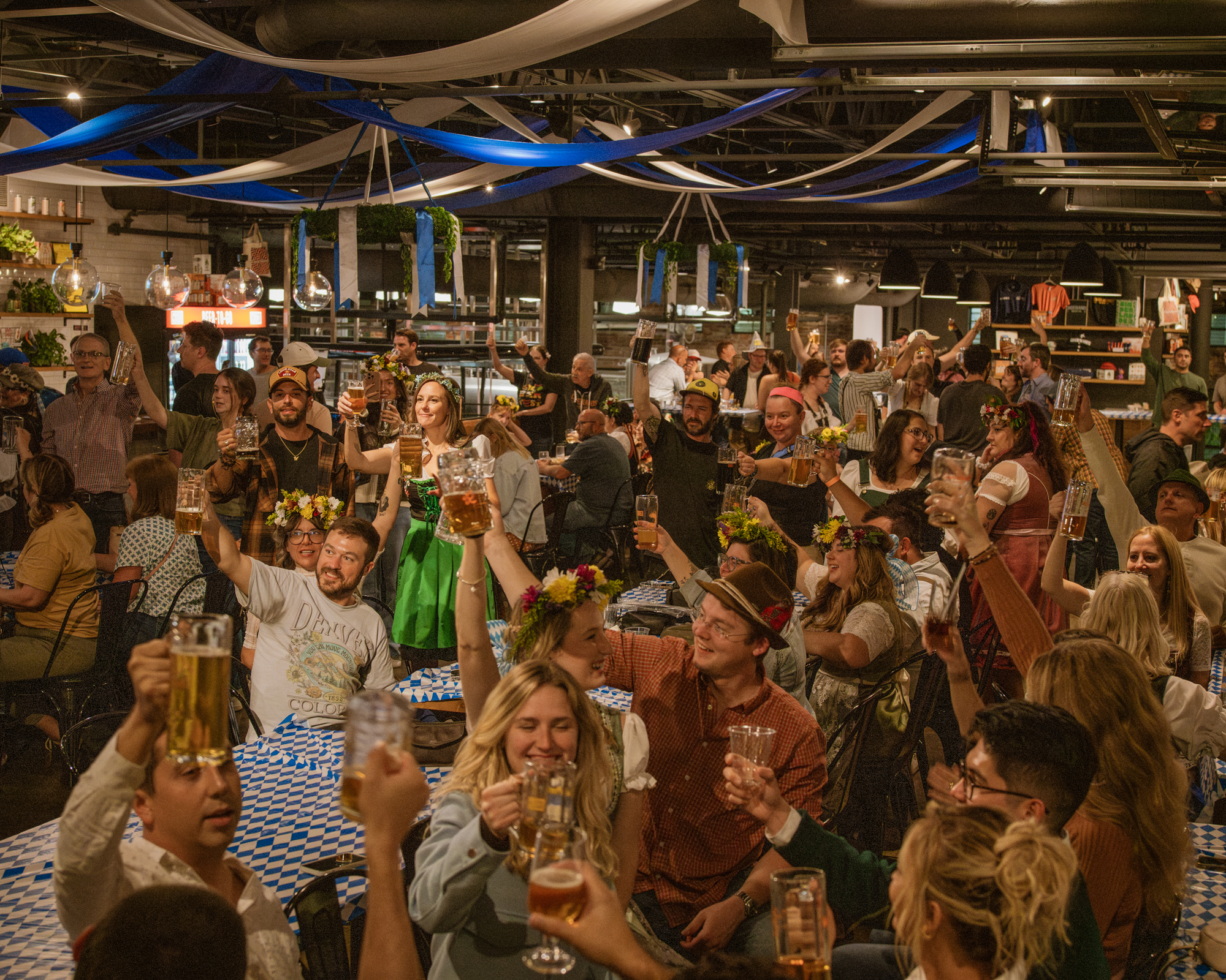 A packed Cinderlands Warehouse at Cinderfest, everyone raising a stein under blue and white streamers